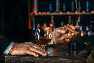 Man's hands with a cigar, elegant glass of brandy on the bar counter. Alcoholic drinks, cognac, whiskey, port, brandy, rum, scotch, bourbon. Vintage wooden table in a pub at night