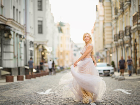 Pretty smiling young woman with long blond hair in elegant flying light dress running along the street