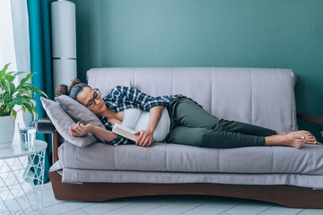 Middle age pregnant woman in eyeglasses wearing checkered shirt sleep lying on grey couch in living room hugging massive book. Daydream, nap, lack of sleeping