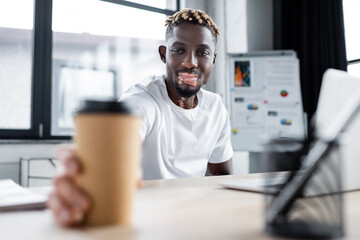 young african american man with vitiligo skin smiling near blurred paper cup in office.