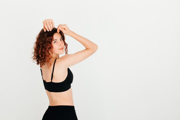 Fototapeta na wymiar Beautiful curly redhead woman posing in black sportswear, turning around to look at the camera, on white studio background