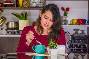 Beautiful latin young brown haired woman in a red sweater sitting in front of table in the dining room, enjoying a hot coffee in an aqua colored mug.