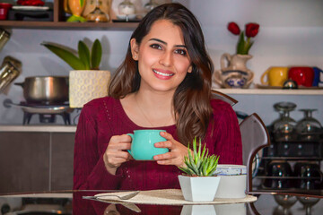 Beautiful latin young brown haired woman in a red sweater sitting in front of table in the dining room, enjoying a hot coffee in an aqua colored mug.