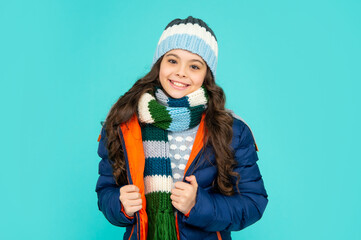 happy kid with curly hair in hat and scarf. teen girl in puffer jacket on blue background