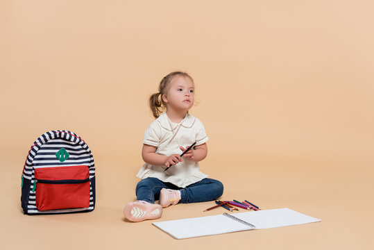 kid with down syndrome sitting near colorful pencils and backpack on beige.