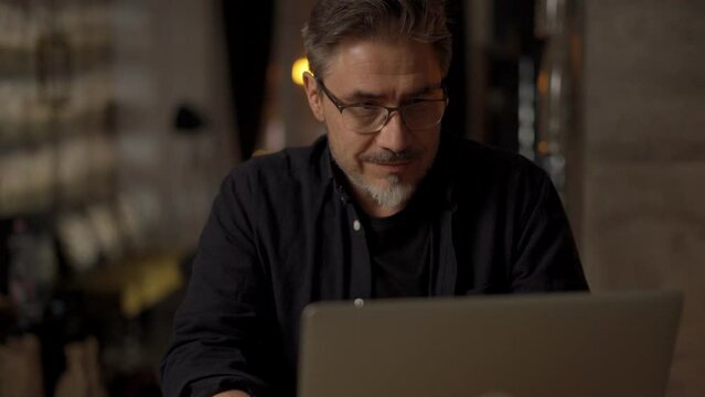 Happy mid adult man working with laptop computer in home office. Businessman smiling, thinking, wearing glasses.