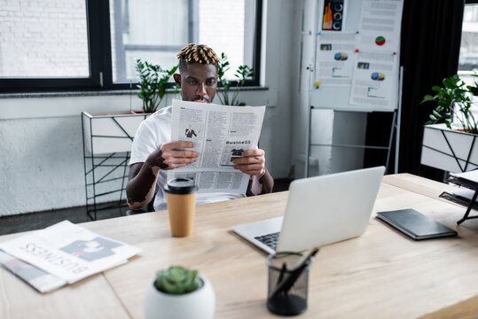 african american man with vitiligo reading newspaper near laptop and blurred flip chart in office.