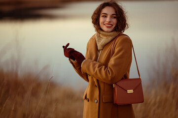 Outdoor autumn portrait of happy smiling fashionable woman wearing trendy outfit: brown coat, turtleneck sweater, suede gloves, with stylish faux leather shoulder bag. Copy, empty space for text