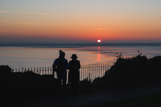 Sunset over the nature reserve of Pewgell Bay. A couple in the foreground in silhouette wearing winter clothes are watching the sunset from the cliff tops in Ramsgate