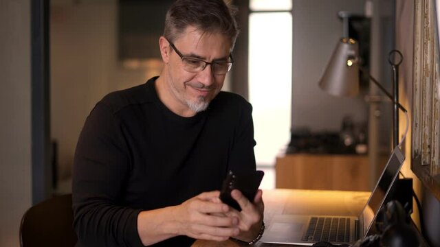 Casual man working with phone and laptop computer in home office, happy, smiling. Mature age, middle age, mid adult man in 50s, white caucasian,