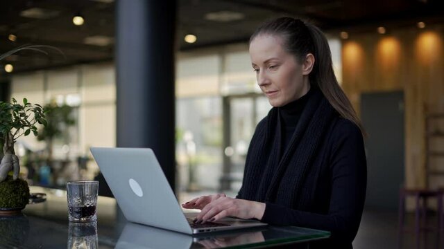 Woman sitting at table in restaurant, cafe or hotel lobby, working with laptop computer. Young authentic businesswoman in business casual, confident happy smiling.