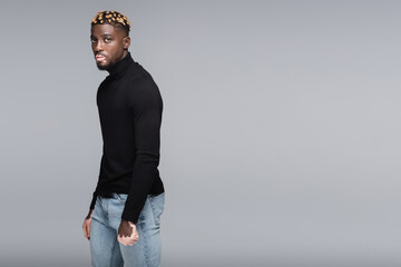 african american man with vitiligo and trendy hairstyle posing in jeans and black turtleneck isolated on grey.