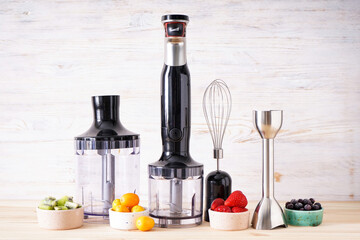 Black plastic electrical hand blender and accessories with fruit on a wooden background.