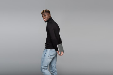 african american man with vitiligo looking at camera while holding laptop behind back isolated on...