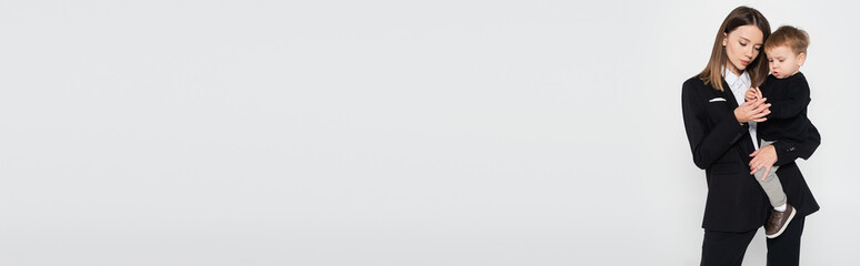 stylish mother in suit holding in arms toddler son isolated on grey, banner.