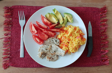 Healthy scrambled egg breakfast with tomatoes and avocadoes on a white plate, set on a red placemat on a wooden table