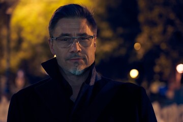 Fototapeta premium Mid adult man on the street at night wearing black coat. Mature age, middle age, mid adult man in 50s, serious look. Copy space.