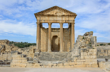 Naklejka premium Temple of Jupiter, forum and ancient roman ruins of Dougga in Tunisia, Africa in the sunny afternoon. Blue sky with clouds, old yellow, grey and brown stone walls and columns