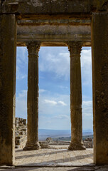 Fototapeta premium View out of the temple of Jupiter in the ancient roman ruined city of Dougga in Tunisia, Africa in the sunny afternoon. Blue sky with clouds, old yellow, grey and brown stone walls and columns
