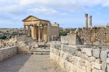 Obraz premium Temple of Jupiter, forum and ancient roman ruins of Dougga in Tunisia, Africa in the sunny afternoon. Blue sky with clouds, old yellow, grey and brown stone walls and columns, paved street
