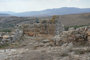 Ruins of the ancient roman city of Dougga in modern Tunisia, Africa. Light sky with clouds, brown, grey and yellow stone walls, green olive trees, mountains on the horizon