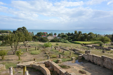 Blue sky with clouds above the antique Roman city of Carthage. Yellow, grey and brown ruined stone walls and columns, red Tunisian flags, green grass and trees, blue waters of the Mediterranean sea.