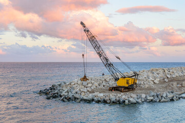 Heavy crane builds a new stone harbour on a sea shore. Blue evening sunset sky with rose clouds,...
