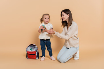 girl with down syndrome holding small globe near happy mother and backpack on beige.