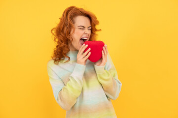 hungry redhead woman biting red heart on yellow background. love