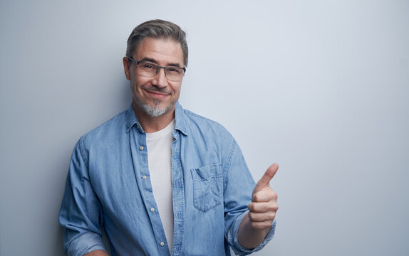 Portrait of mature age, middle age, mid adult casual man in 50s, happy confident smile, standing at wall wearing glasses showing thumb up. White background, copy space.