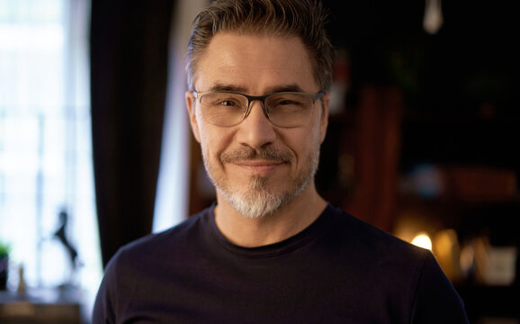 Portrait of mature age, middle age, mid adult casual man in glasses, confident happy smiling. Dark cosy room, copy space.
