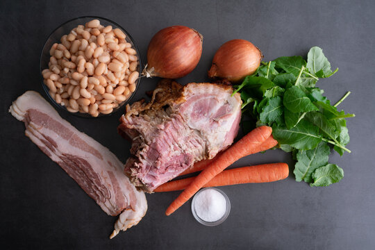 Ham Bone Soup with White Beans and Kale  Ingredients: Overhead view of hambone, bacon, and vegetables on a dark background