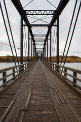 Obraz premium Steel trestle bridge with wooden deck over a large river in Newfoundland. The bridge is for foot traffic and ATV usage. The sky is clear blue and land and houses can be seen in the background. 