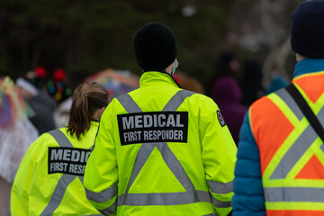 Medical first responders walking along a road wearing black wool stocking caps, yellow reflective coats with the medical first responder in grey letters and across. The EMT is carrying a first aid kit