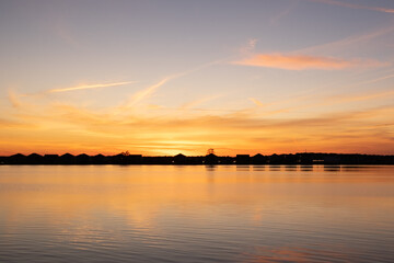 A bright orange sunset over the ocean with wispy clouds. The sky has some dark clouds and the sun is just going down over the horizon. There are water ripples in the foreground of the photo.
