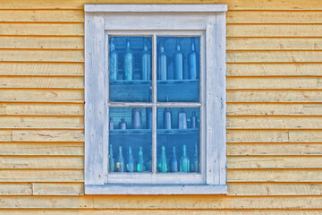 The top corner of a yellow wooden building. There's a double hung window with white trim. The edge of the building has white trim. The window has four colorful shelves lined with vintage bottles.  