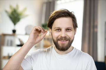 Cheerful bearded young man holding cotton swab sticks for ear cleaning, looking at camera, posing on the background of modern room. Skin care, ear hygiene, healthcare cosmetic procedures concept