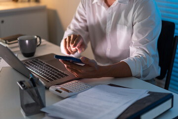 Overwork Concept The office man who sits at his desk holding his smartphone for searching some...