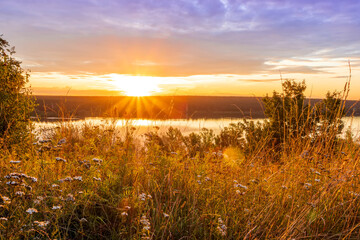 Amazing view at beautiful sunset or sunrise on a shiny lake from hill with green bushes, golden sun rays, calm water ,deep blue cloudy sky and forest on a background