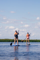 Low angle of older ladies on sup boards both moving oars in the same way on blue lake with ripples with green reeds in background wearing swimming suits. Active lifestyle for older people. 
