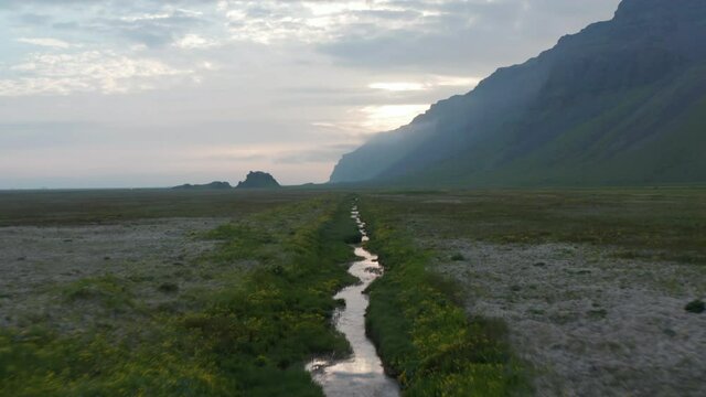 Backwards fly above small creek flowing through flat meadow. Steep rocky escarpment aside and sun behind clouds. Revealing standing SUV car. Iceland