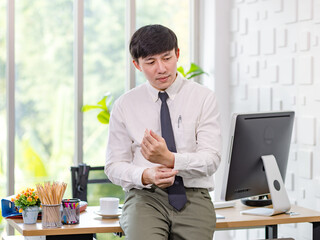 Asian young handsome professional successful male businessman employee in formal business shirt adjusting necktie sitting leaning on working desk full of computer monitor stationery and coffee cup