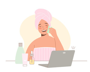 Concept of online beauty treatments. View online classes on laptop. Young woman with towel on her head takes care of her face. She uses jade gouache scraper for massage.