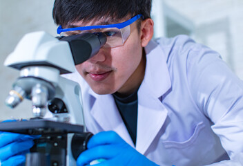 Closeup facial shot of Asian young professional male scientist in white lab coat wearing safety protection goggles glasses using microscope lens looking zooming at microbiology sample on glass plate