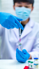 Closeup shot of syringe needle sucking coronavirus covid19 vaccine from glass vial bottle for injection by Asian professional male scientist in white lab coat wearing face mask in blurred background