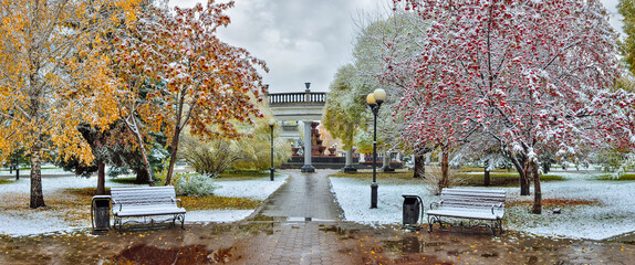 Cozy corner of autumn city park after first snowfall - late autumn or early winter landscape. Colorful fall foliage, red berries of rowan and wild apple trees, benches with white snow covered - fairy 