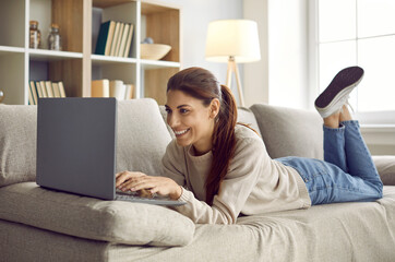 Happy woman using her laptop computer. Smiling young girl lying on a comfy sofa at home, using her modern notebook PC, working remotely or studying a new online course