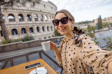 Woman having fun taking selfie with coffee at outdoor cafe on background of coliseum, the most...