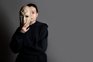 Hiding behind a mask, a young woman in a dark hoodie hides her face with a mask, the concept of self-doubt. Portrait in the studio on a dark gray background.