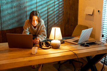 Young focused woman using smart phone while sitting at cozy home office during the nighttime. Concept of online work from home. Idea of cozy and style workplace